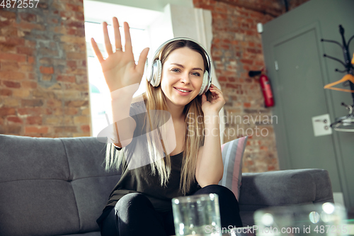 Image of Young woman wearing wireless headphones gesturing during a video conference in the living room. Waving