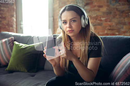 Image of Young woman wearing wireless headphones gesturing during a video conference in the living room. Pointing on phone