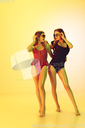 Image of Beautiful girls in fashionable swimsuits isolated on yellow studio background in neon light. Summer, resort, fashion and weekend concept.