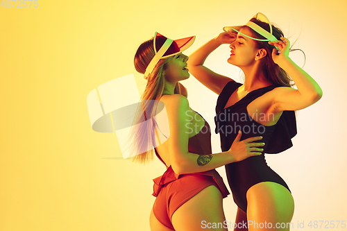 Image of Beautiful girls in fashionable swimsuits isolated on yellow studio background in neon light. Summer, resort, fashion and weekend concept. Wearing stylish red beach hats.