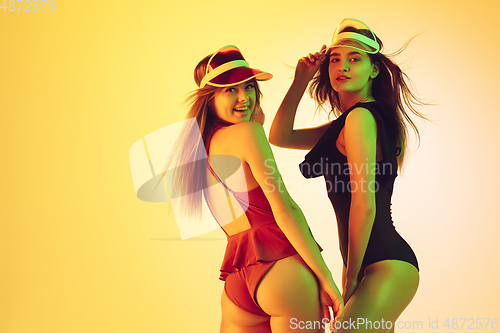 Image of Beautiful girls in fashionable swimsuits isolated on yellow studio background in neon light. Summer, resort, fashion and weekend concept. Wearing stylish red beach hats.