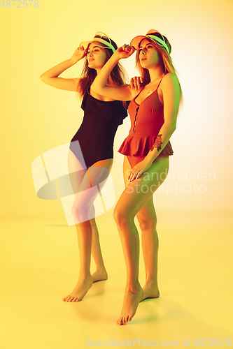 Image of Beautiful girls in fashionable swimsuits isolated on yellow studio background in neon light. Summer, resort, fashion and weekend concept.