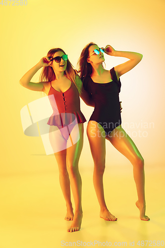 Image of Beautiful girls in fashionable swimsuits isolated on yellow studio background in neon light. Summer, resort, fashion and weekend concept.