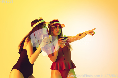 Image of Beautiful girls in fashionable swimsuits isolated on yellow studio background in neon light. Summer, resort, fashion and weekend concept. Drinking cocktails.
