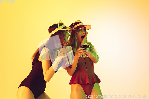 Image of Beautiful girls in fashionable swimsuits isolated on yellow studio background in neon light. Summer, resort, fashion and weekend concept. Drinking cocktails.