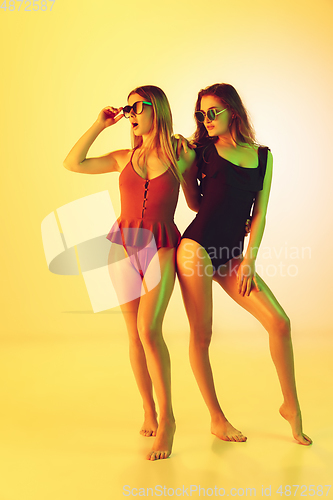 Image of Beautiful girls in fashionable swimsuits isolated on yellow studio background in neon light. Summer, resort, fashion and weekend concept.