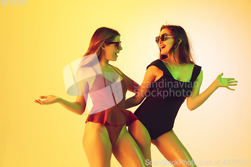 Image of Beautiful girls in fashionable swimsuits isolated on yellow studio background in neon light. Summer, resort, fashion and weekend concept. Dancing.