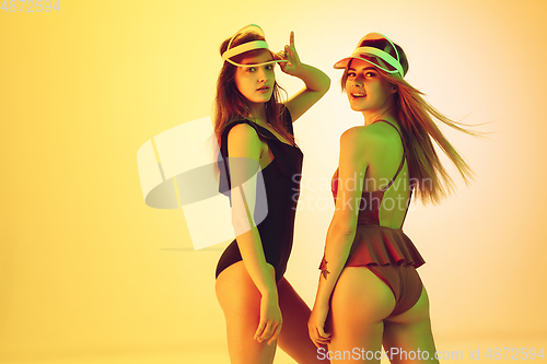 Image of Beautiful girls in fashionable swimsuits isolated on yellow studio background in neon light. Summer, resort, fashion and weekend concept. Wearing stylish red beach hats.