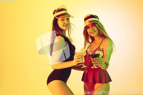 Image of Beautiful girls in fashionable swimsuits isolated on yellow studio background in neon light. Summer, resort, fashion and weekend concept. Drinking cocktails.