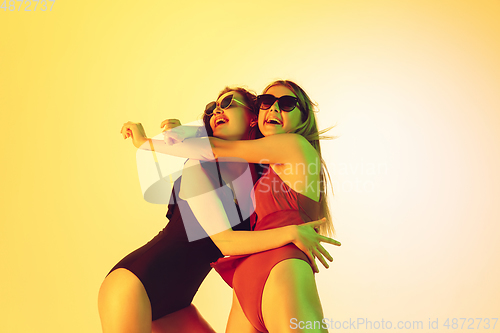 Image of Beautiful girls in fashionable swimsuits isolated on yellow studio background in neon light. Summer, resort, fashion and weekend concept. Dancing.