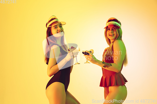 Image of Beautiful girls in fashionable swimsuits isolated on yellow studio background in neon light. Summer, resort, fashion and weekend concept. Drinking cocktails.