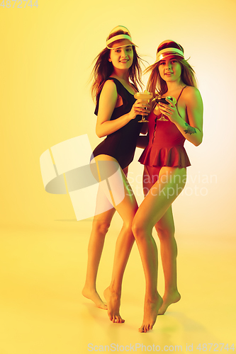 Image of Beautiful girls in fashionable swimsuits isolated on yellow studio background in neon light. Summer, resort, fashion and weekend concept. Drinking cocktails.