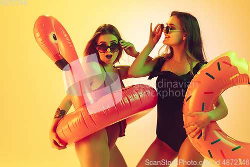 Image of Beautiful girls in fashionable swimsuits isolated on yellow studio background in neon light. Summer, resort, fashion and weekend concept. Posing in rubber donut and flamingo with bright emotions.