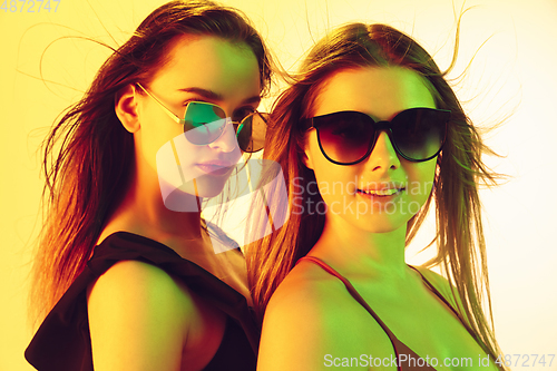 Image of Close up beautiful girls in fashionable swimsuits and eyewear isolated on yellow studio background in neon light. Summer, resort, fashion and weekend concept.