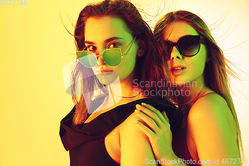 Image of Close up beautiful girls in fashionable swimsuits and eyewear isolated on yellow studio background in neon light. Summer, resort, fashion and weekend concept.