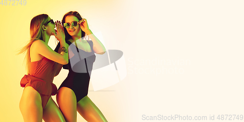 Image of Beautiful girls in fashionable swimsuits isolated on yellow studio background in neon light. Summer, resort, fashion and weekend concept. Flyer with copyspace.