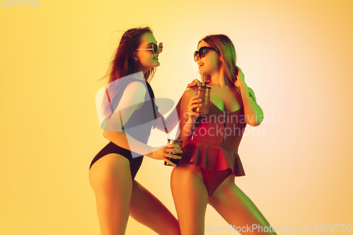 Image of Beautiful girls in fashionable swimsuits isolated on yellow studio background in neon light. Summer, resort, fashion and weekend concept. Dancing with cocktails.
