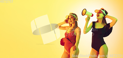 Image of Beautiful girls in fashionable swimsuits and eyewear, headphones isolated on yellow studio background in neon light. Summer, resort, fashion and weekend concept. Calling with megaphone. Flyer.