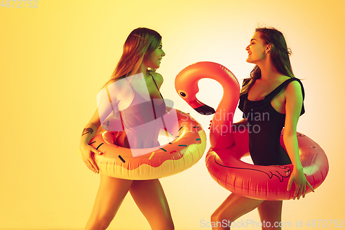 Image of Beautiful girls in fashionable swimsuits isolated on yellow studio background in neon light. Summer, resort, fashion and weekend concept. Posing in rubber donut and flamingo with bright emotions.