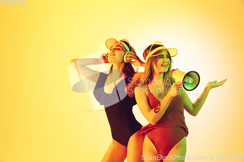 Image of Beautiful girls in fashionable swimsuits and eyewear, headphones isolated on yellow studio background in neon light. Summer, resort, fashion and weekend concept. Calling with megaphone.