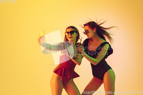 Image of Beautiful girls in fashionable swimsuits isolated on yellow studio background in neon light. Summer, resort, fashion and weekend concept. Taking selfie with cocktails.