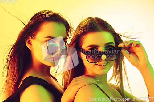 Image of Close up beautiful girls in fashionable swimsuits and eyewear isolated on yellow studio background in neon light. Summer, resort, fashion and weekend concept.