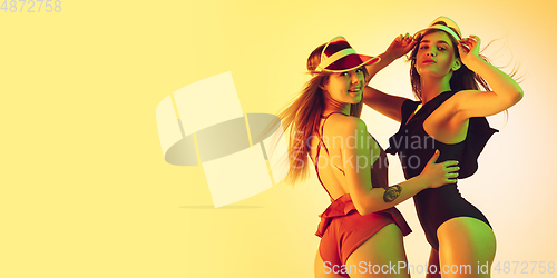 Image of Beautiful girls in fashionable swimsuits isolated on yellow studio background in neon light. Summer, resort, fashion and weekend concept. Wearing stylish red beach hats. Flyer with copyspace.