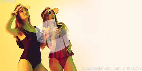 Image of Beautiful girls in fashionable swimsuits isolated on yellow studio background in neon light. Summer, resort, fashion and weekend concept. Dancing with cocktails. Flyer with copyspace.