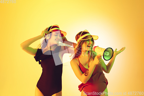 Image of Beautiful girls in fashionable swimsuits and eyewear, headphones isolated on yellow studio background in neon light. Summer, resort, fashion and weekend concept. Calling with megaphone.