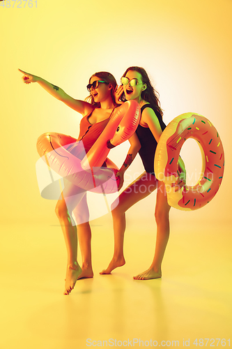 Image of Beautiful girls in fashionable swimsuits isolated on yellow studio background in neon light. Summer, resort, fashion and weekend concept. Posing in rubber donut and flamingo with bright emotions.