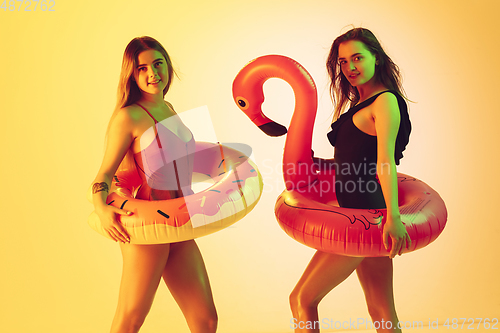Image of Beautiful girls in fashionable swimsuits isolated on yellow studio background in neon light. Summer, resort, fashion and weekend concept. Posing in rubber donut and flamingo with bright emotions.