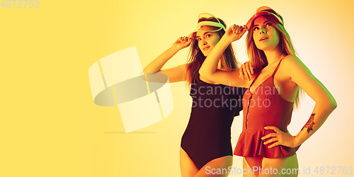 Image of Beautiful girls in fashionable swimsuits isolated on yellow studio background in neon light. Summer, resort, fashion and weekend concept. Wearing stylish red beach hats. Flyer with copyspace.