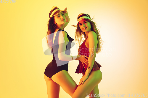 Image of Beautiful girls in fashionable swimsuits isolated on yellow studio background in neon light. Summer, resort, fashion and weekend concept. Wearing stylish red beach hats.