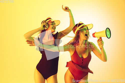 Image of Beautiful girls in fashionable swimsuits and eyewear, headphones isolated on yellow studio background in neon light. Summer, resort, fashion and weekend concept. Calling with megaphone.