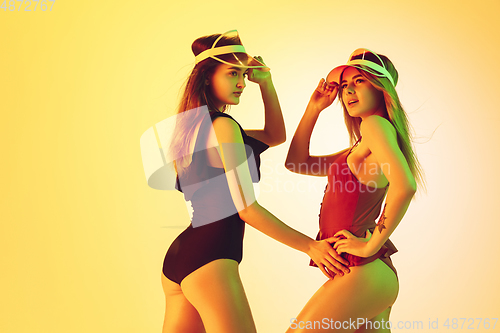 Image of Beautiful girls in fashionable swimsuits isolated on yellow studio background in neon light. Summer, resort, fashion and weekend concept. Wearing stylish red beach hats.