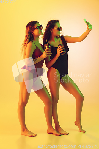 Image of Beautiful girls in fashionable swimsuits isolated on yellow studio background in neon light. Summer, resort, fashion and weekend concept. Taking selfie with cocktails.