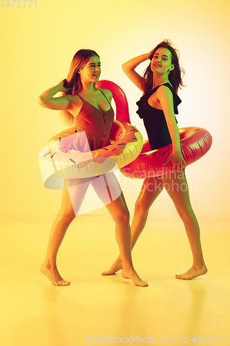 Image of Beautiful girls in fashionable swimsuits isolated on yellow studio background in neon light. Summer, resort, fashion and weekend concept. Posing in rubber donut and flamingo with bright emotions.