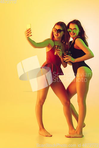 Image of Beautiful girls in fashionable swimsuits isolated on yellow studio background in neon light. Summer, resort, fashion and weekend concept. Taking selfie with cocktails.