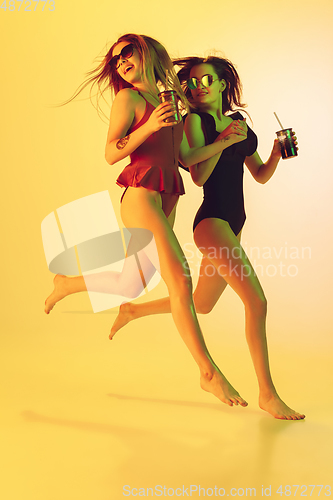 Image of Beautiful girls in fashionable swimsuits isolated on yellow studio background in neon light. Summer, resort, fashion and weekend concept. With cocktails on the run.