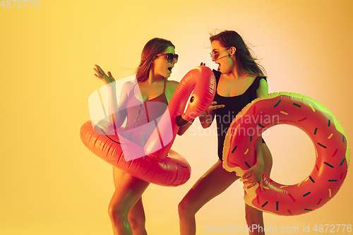 Image of Beautiful girls in fashionable swimsuits isolated on yellow studio background in neon light. Summer, resort, fashion and weekend concept. Posing in rubber donut and flamingo with bright emotions.