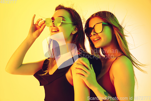 Image of Close up beautiful girls in fashionable swimsuits and eyewear isolated on yellow studio background in neon light. Summer, resort, fashion and weekend concept.