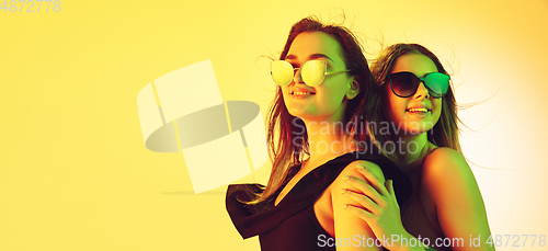 Image of Close up beautiful girls in fashionable swimsuits and eyewear isolated on yellow studio background in neon light. Summer, resort, fashion and weekend concept. Flyer with copyspace.