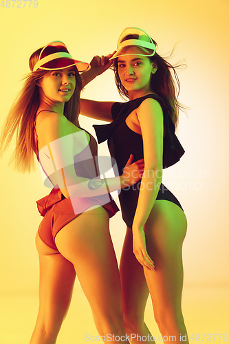 Image of Beautiful girls in fashionable swimsuits isolated on yellow studio background in neon light. Summer, resort, fashion and weekend concept. Wearing stylish red beach hats.