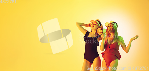 Image of Beautiful girls in fashionable swimsuits and eyewear, headphones isolated on yellow studio background in neon light. Summer, resort, fashion and weekend concept. Calling with megaphone. Flyer.