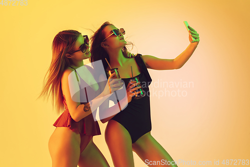 Image of Beautiful girls in fashionable swimsuits isolated on yellow studio background in neon light. Summer, resort, fashion and weekend concept. Taking selfie with cocktails.