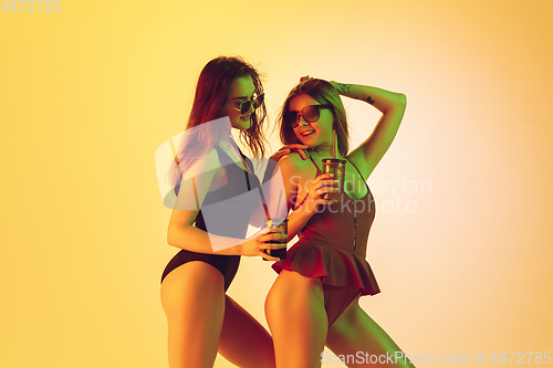 Image of Beautiful girls in fashionable swimsuits isolated on yellow studio background in neon light. Summer, resort, fashion and weekend concept. Dancing with cocktails.