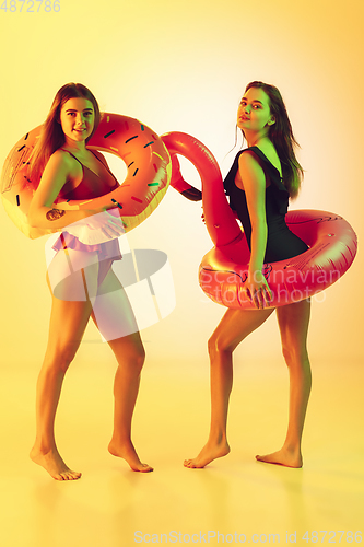 Image of Beautiful girls in fashionable swimsuits isolated on yellow studio background in neon light. Summer, resort, fashion and weekend concept. Posing in rubber donut and flamingo with bright emotions.