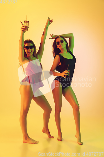 Image of Beautiful girls in fashionable swimsuits isolated on yellow studio background in neon light. Summer, resort, fashion and weekend concept. Dancing with cocktails.