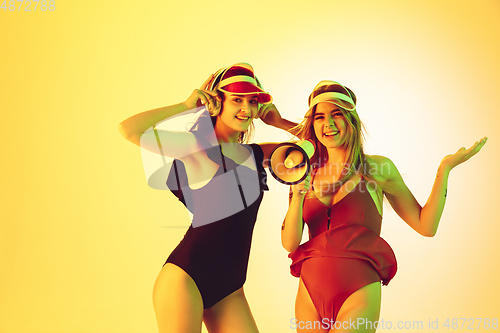 Image of Beautiful girls in fashionable swimsuits and eyewear, headphones isolated on yellow studio background in neon light. Summer, resort, fashion and weekend concept. Calling with megaphone.