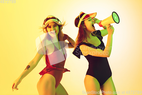 Image of Beautiful girls in fashionable swimsuits and eyewear, headphones isolated on yellow studio background in neon light. Summer, resort, fashion and weekend concept. Calling with megaphone.